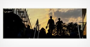 Silhouetted people walk up stairs at sunset, with dramatic clouds and an orange sky in the background, framed by buildings and trees on either side.