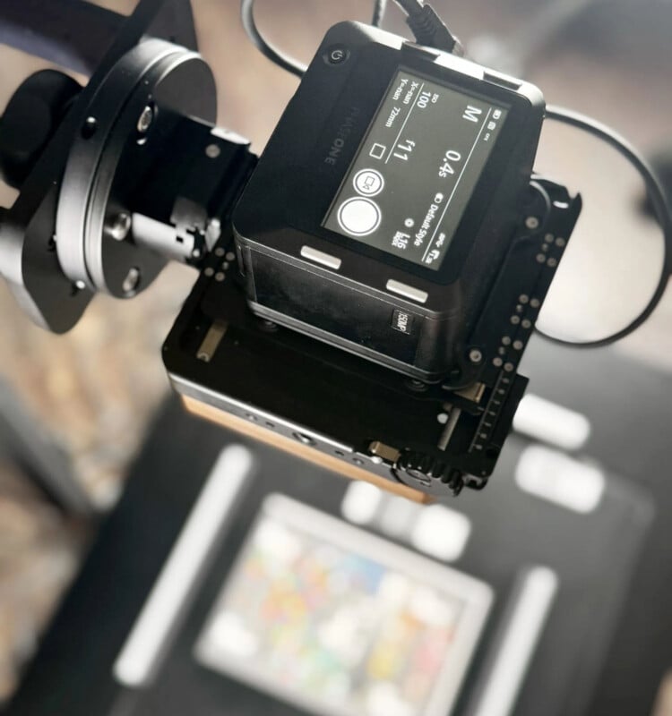 A digital camera mounted on a stand is pointed downward, capturing an image of a colorful, rectangular object placed on a black surface below. The camera's display screen and controls are visible.