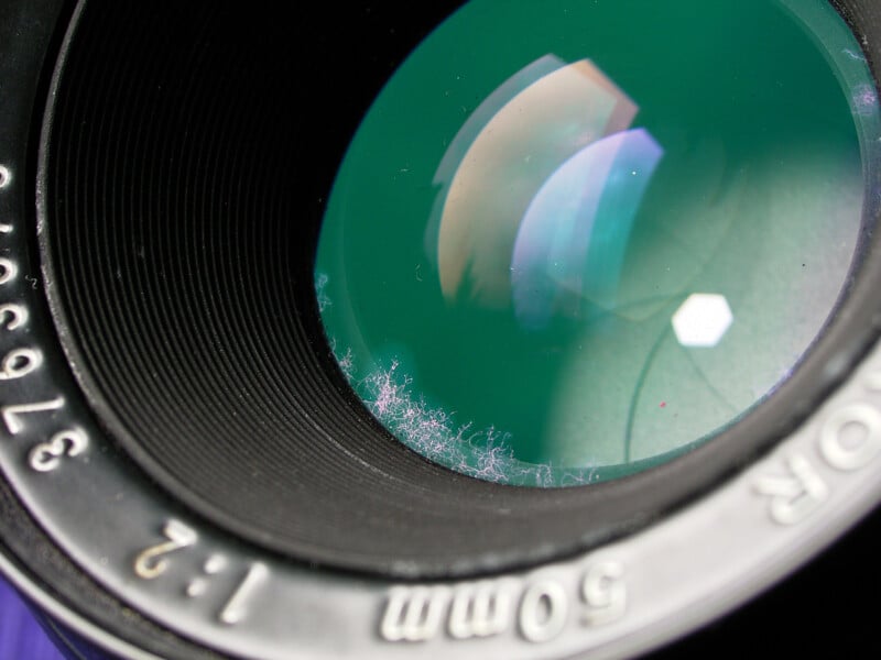Close-up of a camera lens showing dust and lint inside the barrel, with green reflections visible on the lens surface and aperture blades faintly visible.