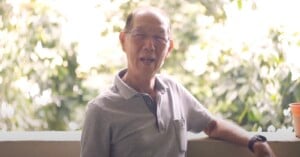 An elderly man with glasses and a grey polo shirt sits outdoors, smiling, with greenery and sunlight in the blurred background. An orange cup is on the right side of the image.