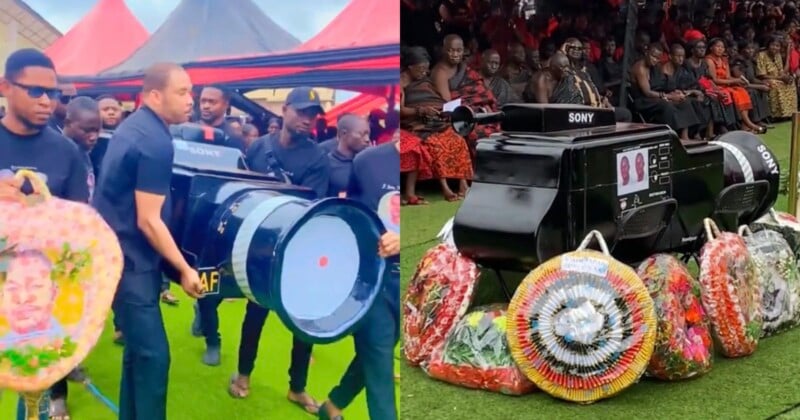 Pallbearers carry and display a coffin shaped like a large Sony camera lens, surrounded by funeral wreaths and mourners dressed in black and red attire at an outdoor ceremony.