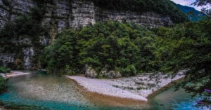 A clear river curves around a gravelly, tree-covered peninsula, with steep, rocky cliffs and dense green forest rising in the background under a cloudy sky.