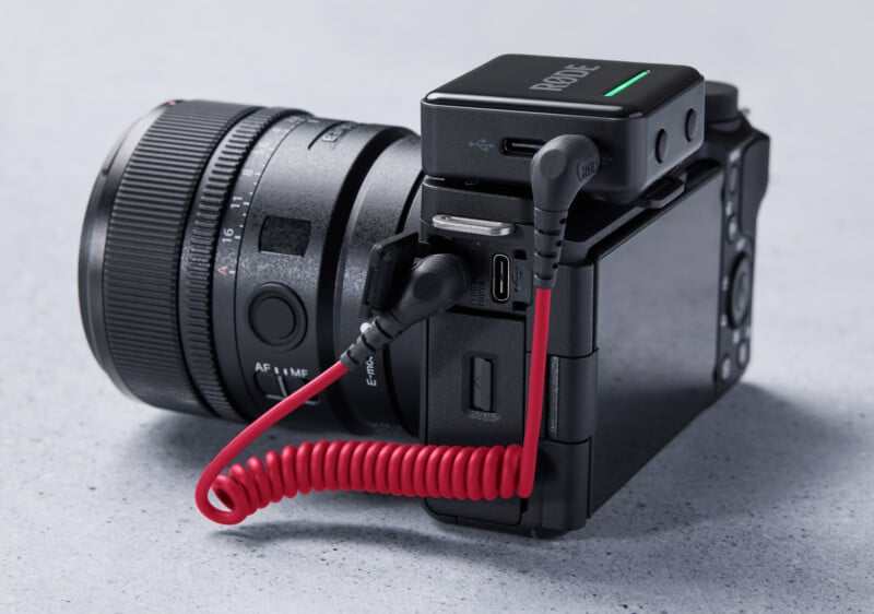 A black digital camera with a lens is connected to a Rode wireless microphone receiver via a red coiled audio cable. The setup is resting on a light gray surface.