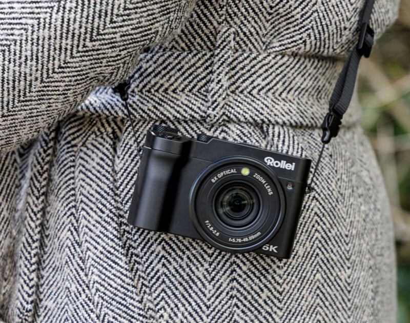 A person wearing a herringbone-patterned coat has a black Rollei digital camera with a strap hanging around their neck.
