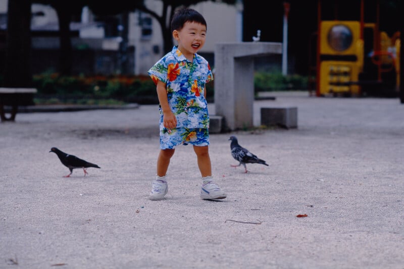 A young child in a colorful floral outfit smiles while walking outdoors in a park, with two pigeons nearby on the ground. Playground equipment and greenery are visible in the background.