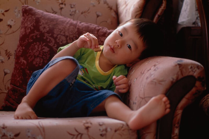 A young child in a green shirt and blue shorts lounges on a patterned couch, resting their head on the armrest and looking thoughtfully at the camera.
