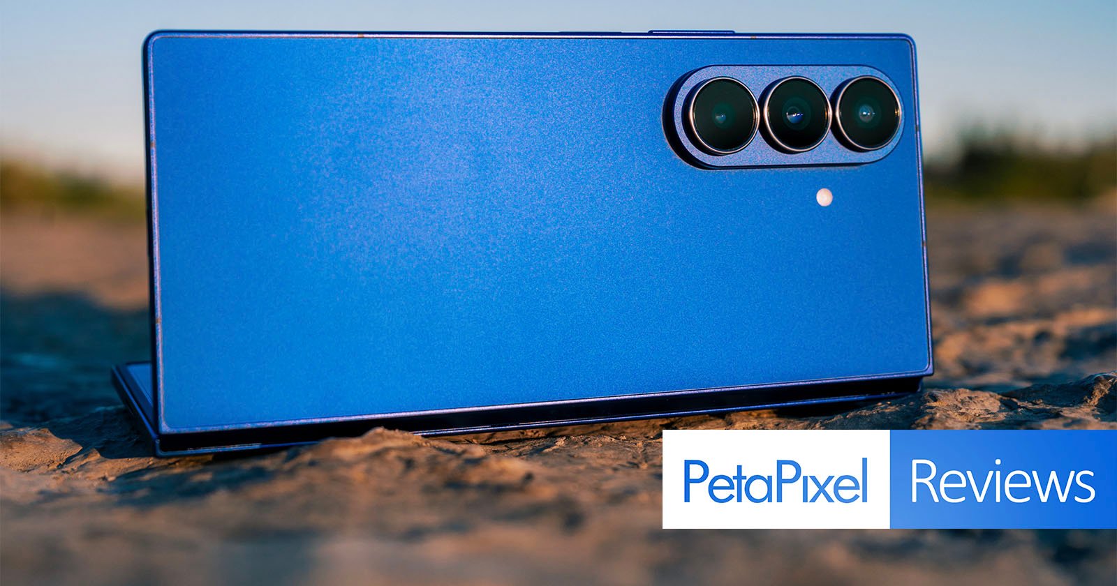 A blue smartphone with three rear cameras laying on a textured outdoor surface, with the "PetaPixel Reviews" logo overlaid in the bottom right corner.