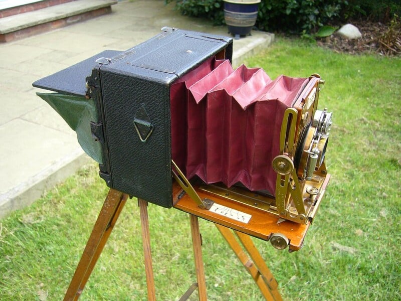 An antique bellows camera with a red accordion-style body sits on a wooden tripod outside on a lawn, with green grass and plants in the background.