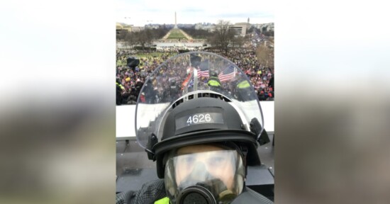 A police officer in riot gear takes a selfie in front of a large crowd gathered near the U.S. Capitol, with American flags visible and the Washington Monument in the background.