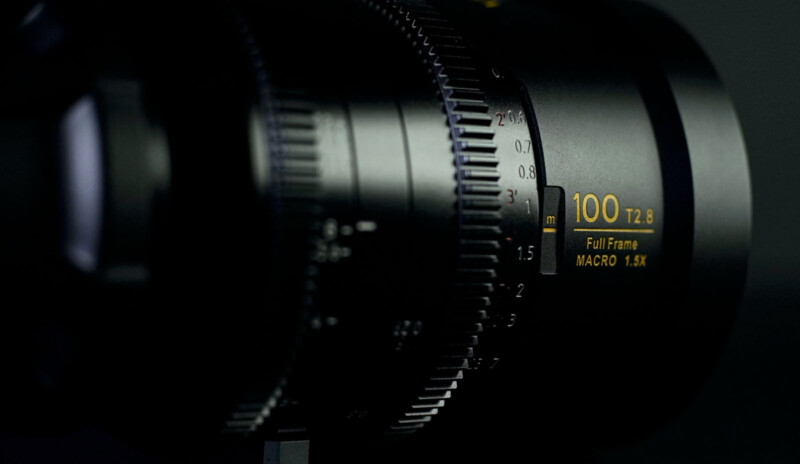 Close-up of a camera lens with focus and aperture markings. The text shows "100 T2.8 Full-Frame MACRO 1.5X" in yellow, and focus distances are visible in white. The lens has a black matte finish.