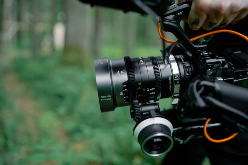 Close-up of a person's tattooed hand holding a professional video camera with a 45mm lens, set against a blurred green forest background. Orange cables are attached to the camera.