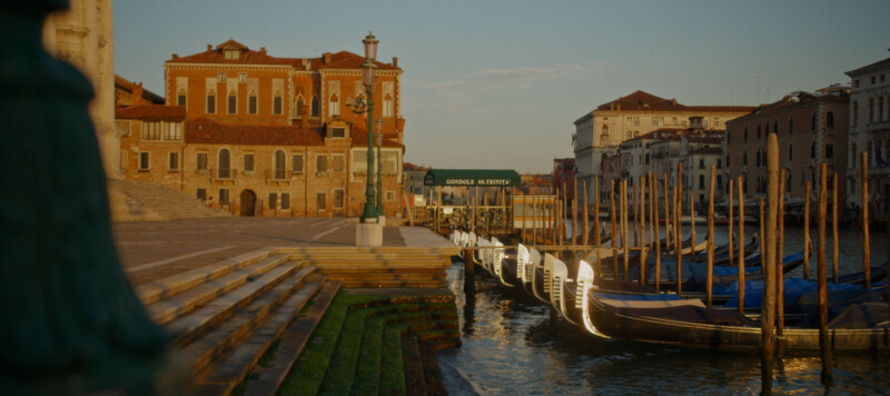 A row of gondolas is moored along the canal in Venice, Italy, with historic buildings and stone steps bathed in warm sunlight, and a sign that reads “Gondole S. S. Trinità.”.