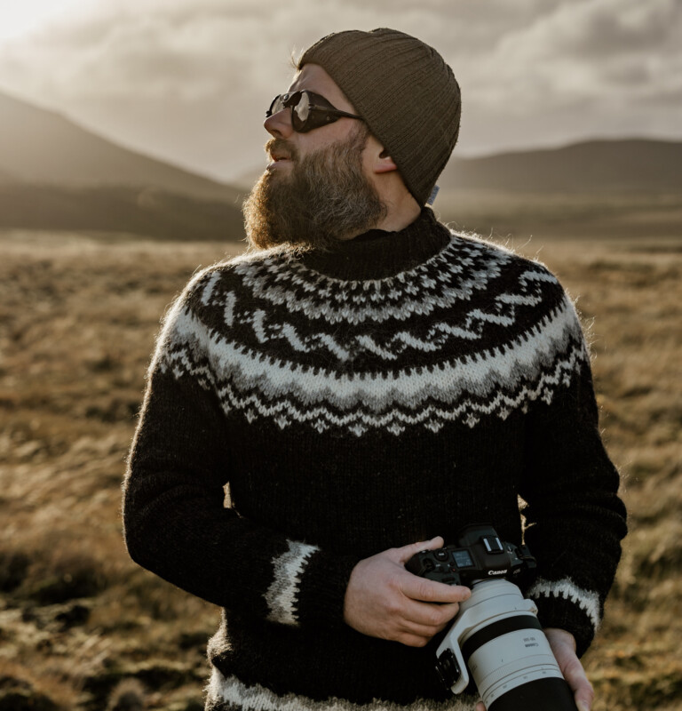 A bearded person in sunglasses and a knit sweater stands outdoors in a grassy, mountainous landscape, holding a camera with a large lens, looking off to the side.