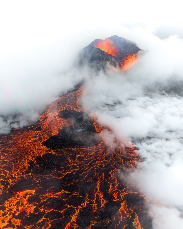 Aerial view of an erupting volcano with bright orange lava flowing down its slopes, surrounded by thick white clouds and dark volcanic rock.