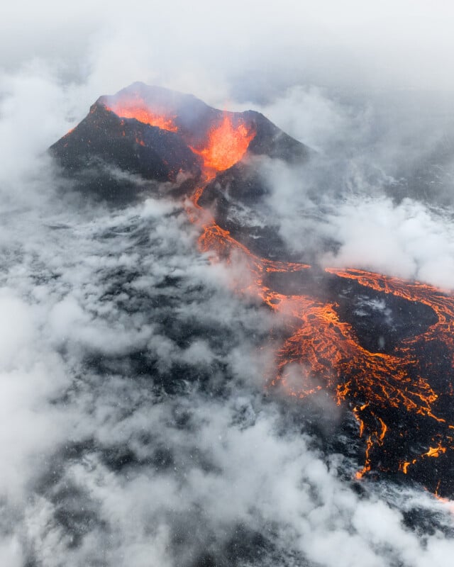 Aerial view of an erupting volcano with bright orange lava flowing down its sides, surrounded by thick clouds of smoke and ash.
