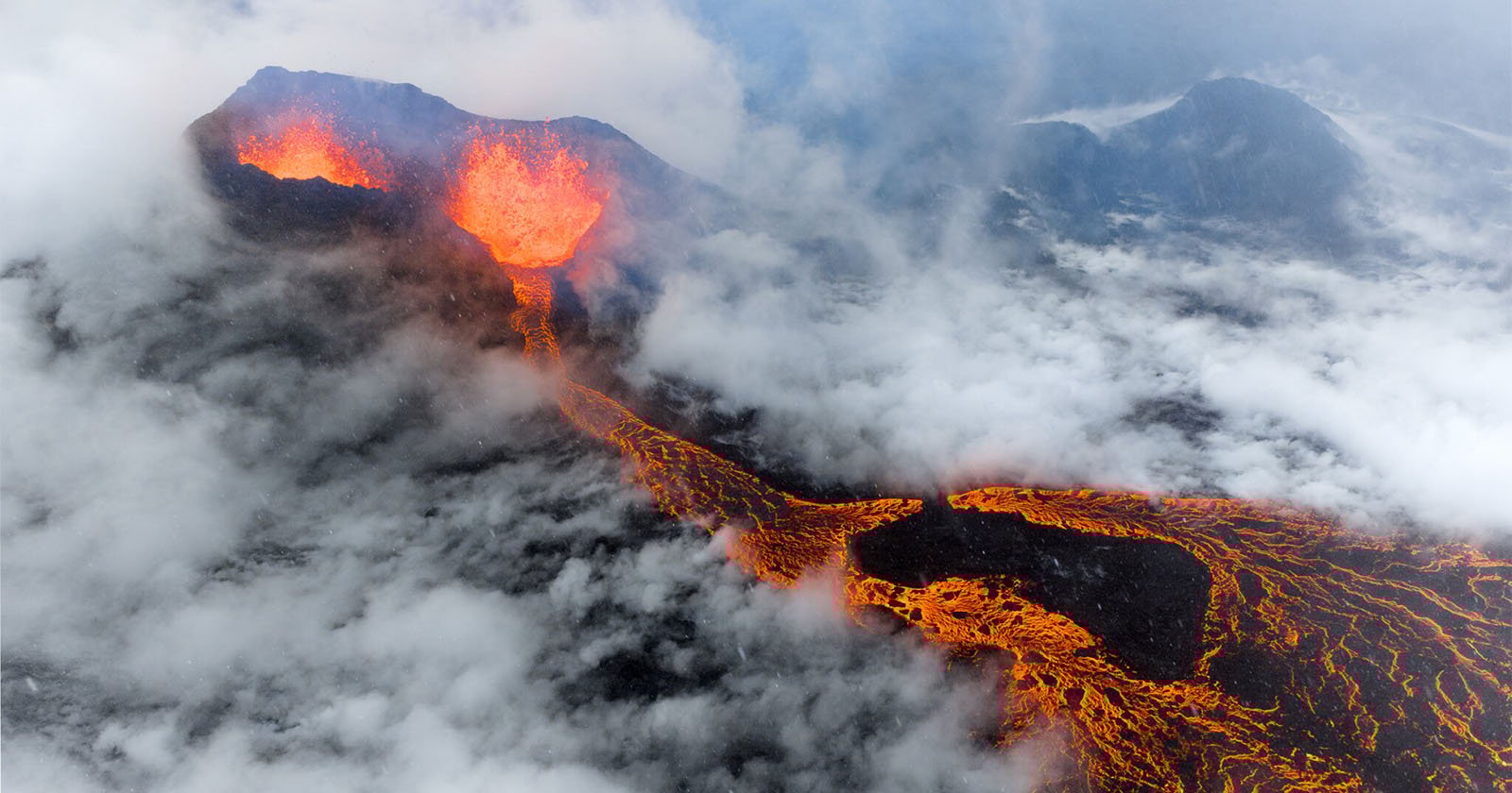 Aerial view of an erupting volcano with bright orange lava flowing down its slopes, surrounded by thick clouds and dark volcanic terrain.