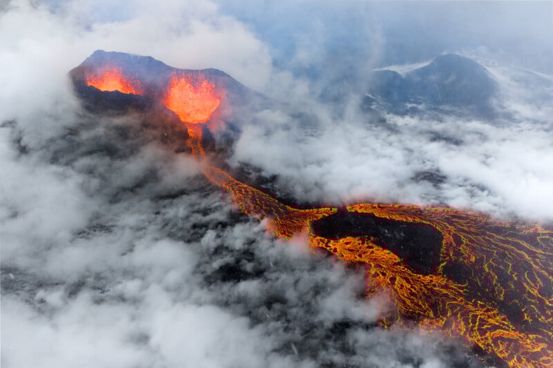 Aerial view of an erupting volcano with bright orange lava flowing down its side, surrounded by dark rocks and thick clouds of white smoke or mist.