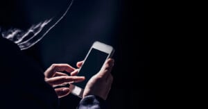 A person wearing a hoodie uses a smartphone in a dark setting, with their face obscured by shadows, creating a mysterious or secretive atmosphere.