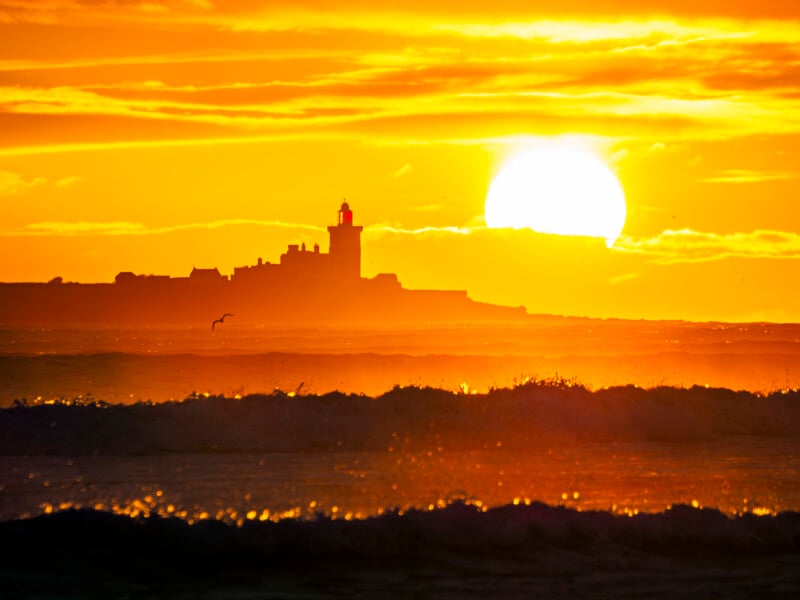 A bright orange sunset over the ocean with waves in the foreground. A silhouette of a lighthouse and buildings appears against the horizon, while the sun glows large in the sky. A bird flies near the lighthouse.