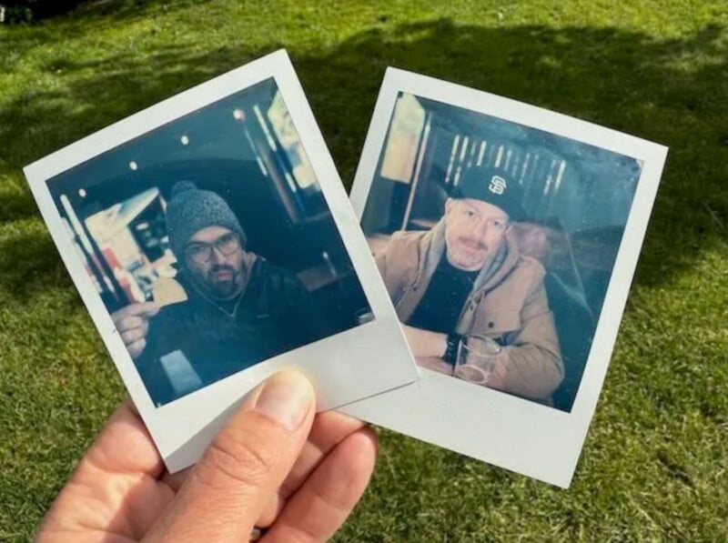 A hand holds two Polaroid photos outdoors on grass, each showing a man in a jacket and beanie or cap, sitting at a table and looking toward the camera.