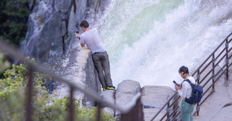 A person in a white shirt leans over a stone barrier near a rushing waterfall, taking photos, while another person with a backpack stands on a walkway, looking at their phone.