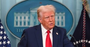 A man in a dark suit and red tie speaks at a podium, with American flags and a blue backdrop of the White House in the background.