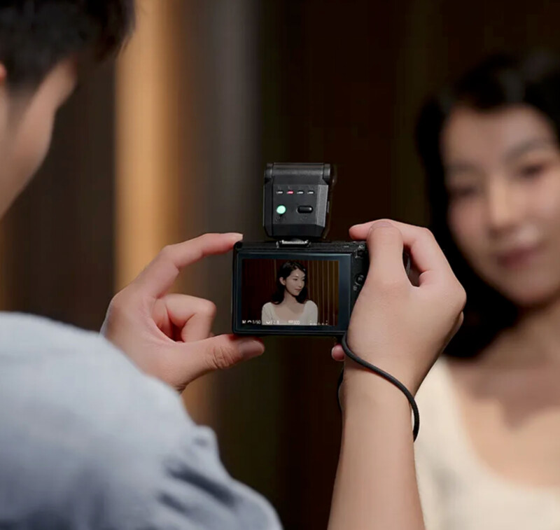 A person holds a digital camera, recording or photographing a woman in a white top. The woman is slightly out of focus in the background but clearly visible on the camera screen.