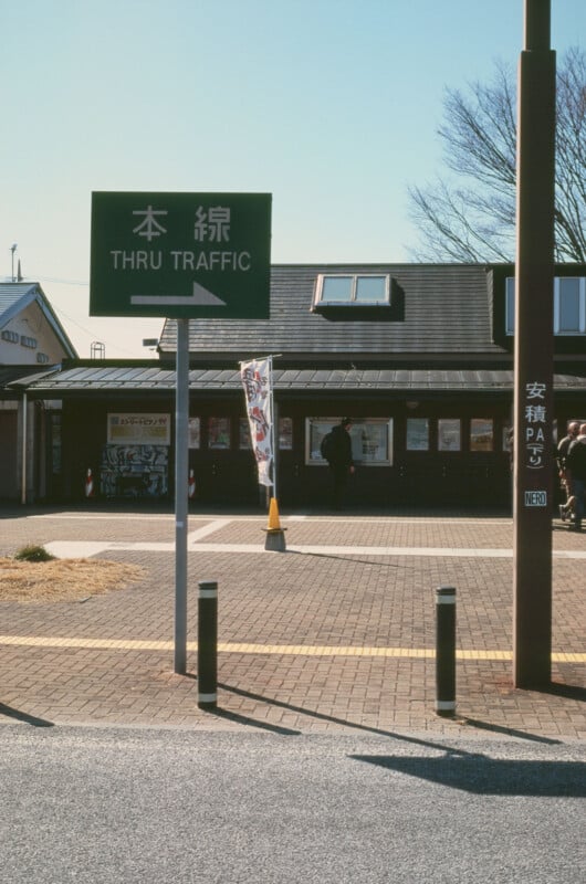 A green "THRU TRAFFIC" sign with Japanese text stands in front of a brick building with large windows, a sloped roof, and a few people near the entrance on a sunny day.