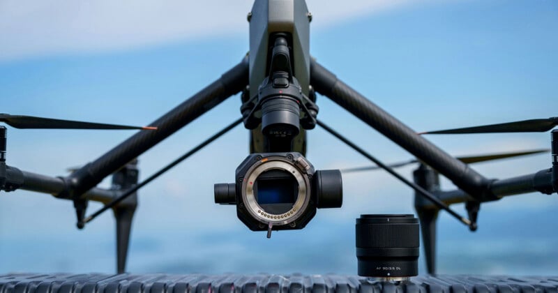Close-up of a professional drone with a camera mount, without the lens attached. A camera lens sits nearby on a textured surface; the background shows a blue sky with clouds.