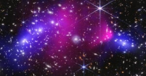 A colorful galaxy cluster with bright stars, glowing purple and blue clouds of gas, and numerous distant galaxies scattered across a dark space background.