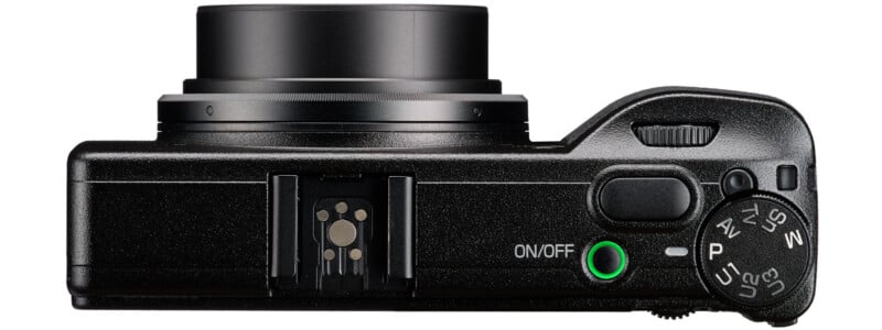Top view of a black digital camera showing the extended lens, mode dial, shutter button, on/off button with a green light, and a hot shoe mount.