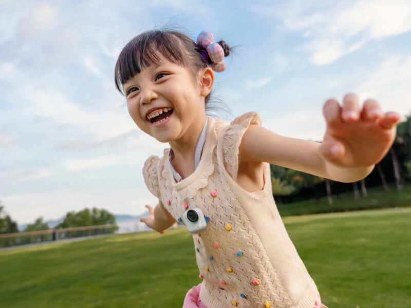 A young girl with a big smile runs outdoors, arms outstretched, wearing a sleeveless sweater with colorful dots and a small camera around her neck. The background shows grass, trees, and a blue sky.