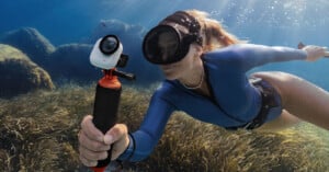 A woman in a blue wetsuit and snorkel mask swims underwater, holding a camera on a stick in front of her, surrounded by rocks and aquatic plants with sunlight streaming from above.
