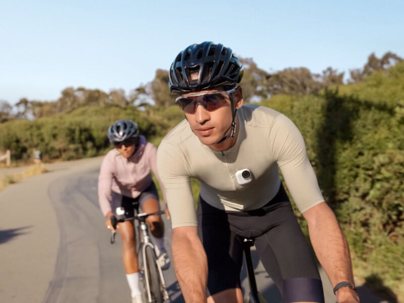Two cyclists ride on a paved path lined with greenery. The man in front wears a helmet, sunglasses, and a light shirt with a camera clipped on, while the woman behind him wears similar cycling gear. The weather is clear and sunny.