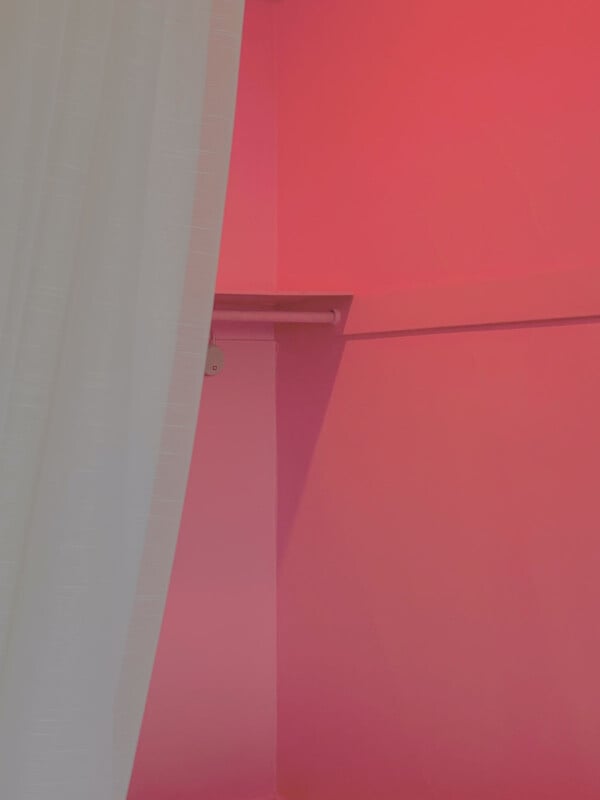 A white curtain partially covers a corner of a room with bright pink walls. A small horizontal shelf or rod is attached to the wall near the top. The space is empty and well-lit.
