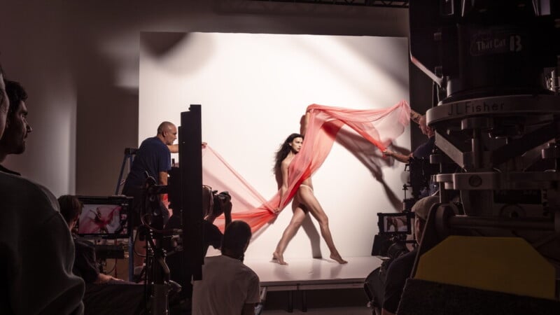 A dancer poses in a photo studio, draped in flowing red fabric held by assistants, as photographers and crew work around her with cameras and lighting equipment.