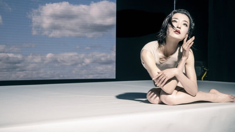 A woman in a pale outfit sits on a white surface, striking an expressive pose. Behind her is a large screen displaying a cloudy sky, contrasting with the dark background.
