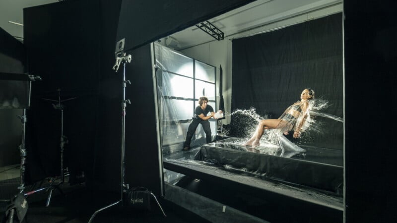 A photographer throws water at a woman sitting on a platform in a studio, capturing the splash as she poses in front of a black backdrop with dramatic lighting and photography equipment visible.
