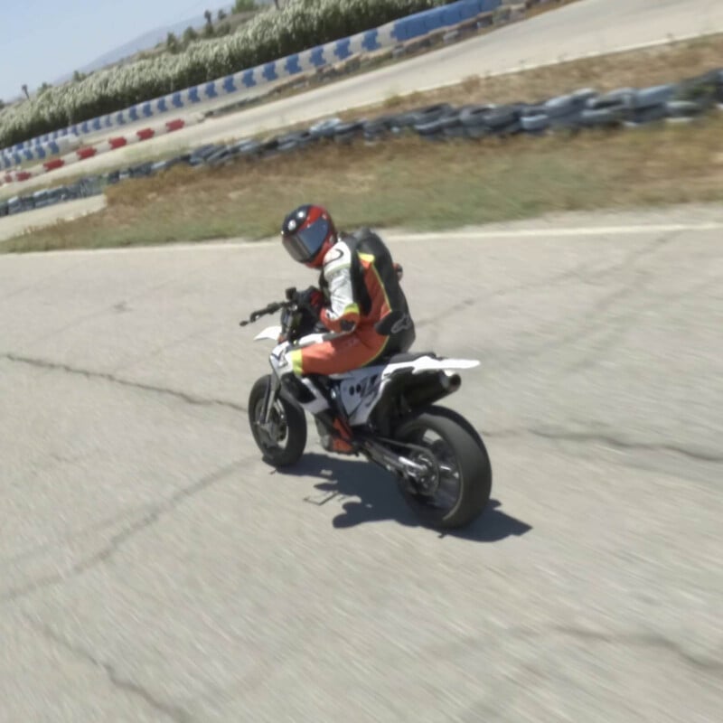 A motorcyclist in a red and black helmet and orange suit leans into a turn on a paved race track, with safety barriers and greenery in the background.