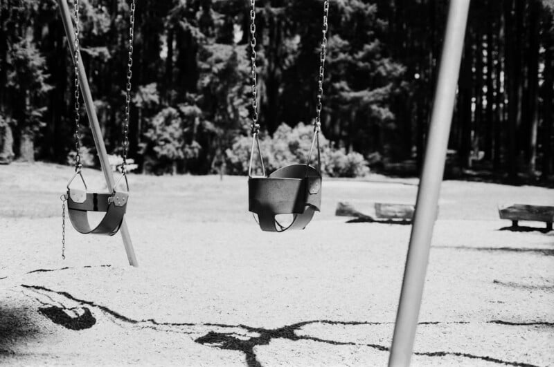 Two empty toddler swings hang from chains at a playground. The area is covered in light gravel or sand with cracks on the ground. Trees and grassy areas are visible in the blurred background. The photo is in black and white.