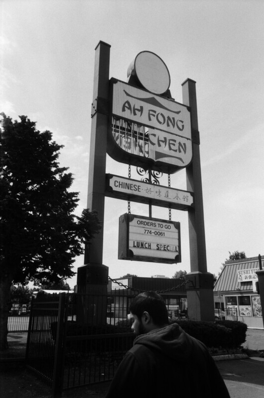 A man walks by a large roadside sign for "Ah Fong Kitchen," advertising Chinese food, orders to go, and lunch specials. Trees and other businesses are visible in the background.