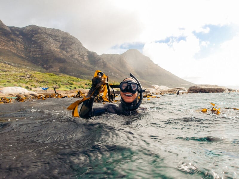 A person in a wetsuit and snorkeling gear smiles while holding seaweed in the ocean, with rocky mountains and a cloudy sky visible in the background.