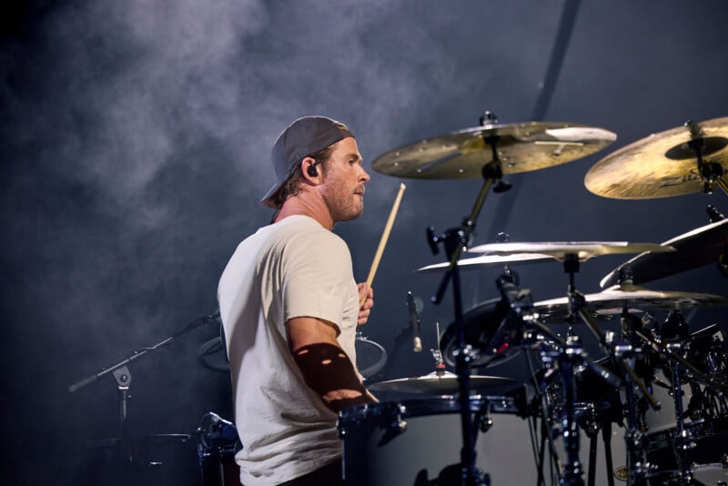 A drummer wearing a backwards cap and white t-shirt plays a drum set on stage, surrounded by cymbals and smoke under dramatic lighting.