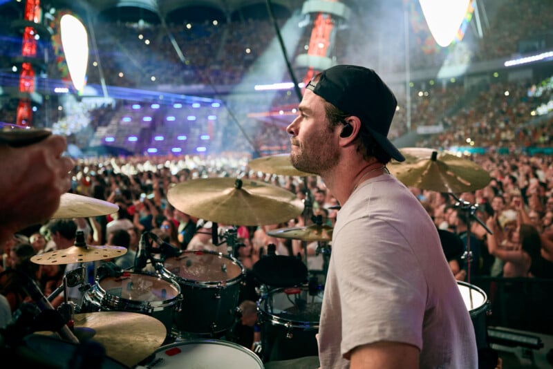 A drummer wearing a backward cap plays drums on stage at a large concert, with a packed, cheering audience and colorful lights in the background.
