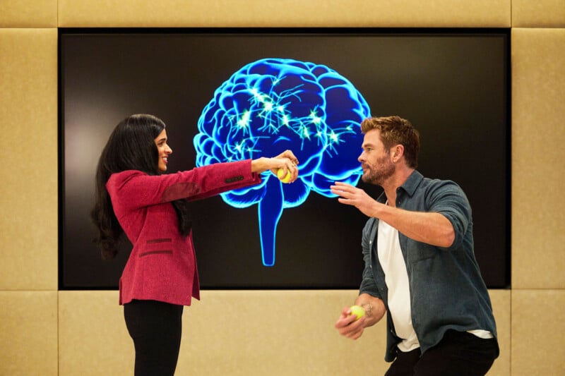 A woman and man interact playfully with yellow objects in front of a large screen displaying a glowing blue brain illustration. The woman extends her arm while the man gestures toward her.