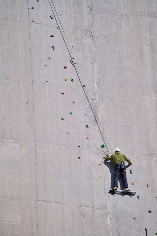 A person wearing a helmet and harness climbs a tall, flat concrete wall using colorful artificial handholds, attached to a safety rope.