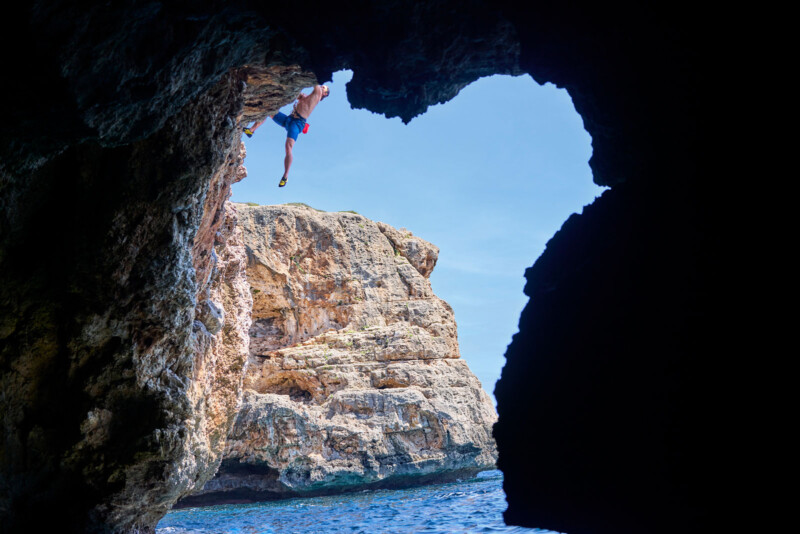 A person climbs the underside of a rocky cliff near the ocean, framed by a cave opening with blue sky and large rock formations visible in the background.