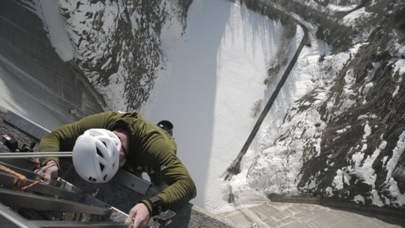 A climber wearing a white helmet and green jacket ascends a snowy, steep dam wall, gripping onto metal rungs with a snowy landscape and winding road far below.