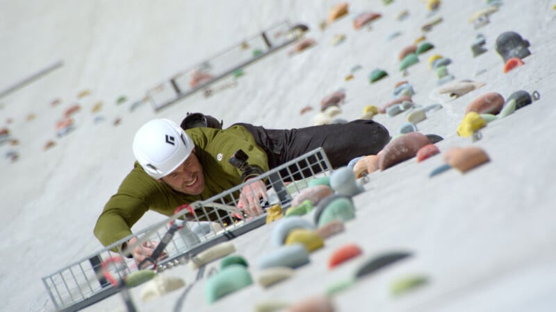 A person wearing a helmet and green jacket climbs an outdoor artificial climbing wall, gripping colorful handholds and using gear, with a determined expression on their face.