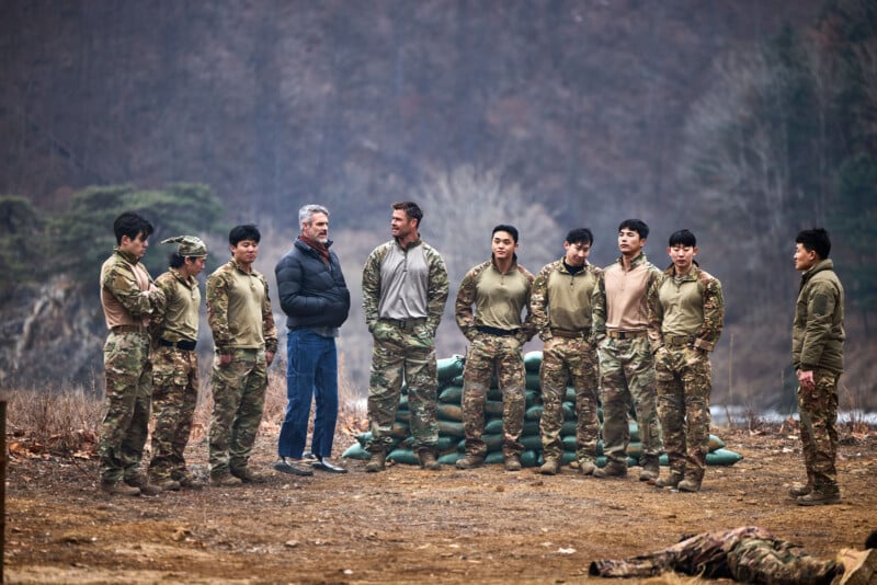A group of soldiers in camouflage pants and tan shirts stand outdoors in a semicircle, listening to two men in civilian clothes. Trees and sandbags are visible in the background.